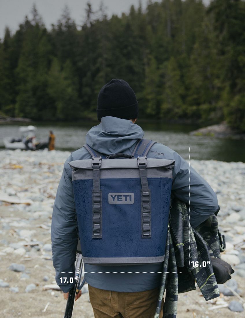 Person with a YETI  cooler backpack standing by a river with trees in the background