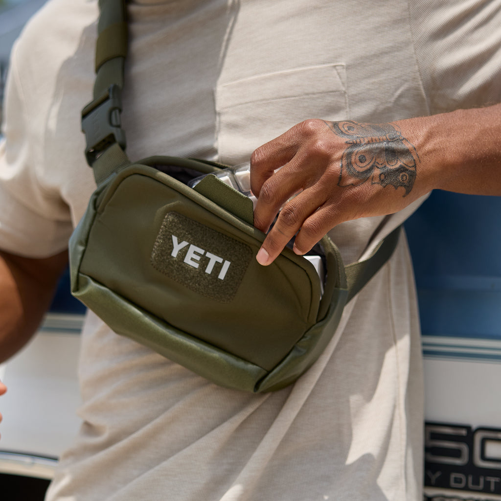 An olive green YETI crossbody bag worn by a person in a light beige shirt, with the person's tattooed hand reaching into the pocket.