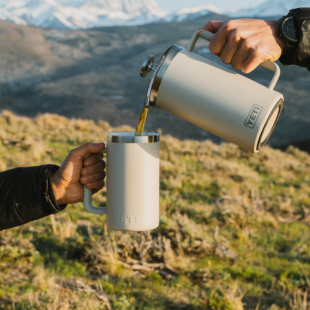 Two hands pouring coffee from a YETI mug into another YETI mug against a mountainous background.