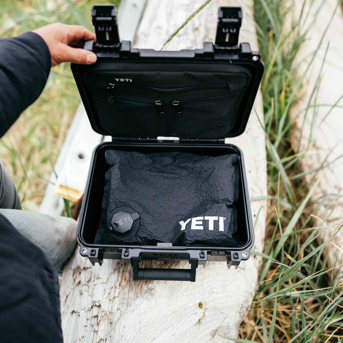 A YETI hard case is open outdoors on a wooden dock, showing a black insulated bag inside with the YETI logo printed in white. A person's hand is visible holding the case open, and grass is in the background.