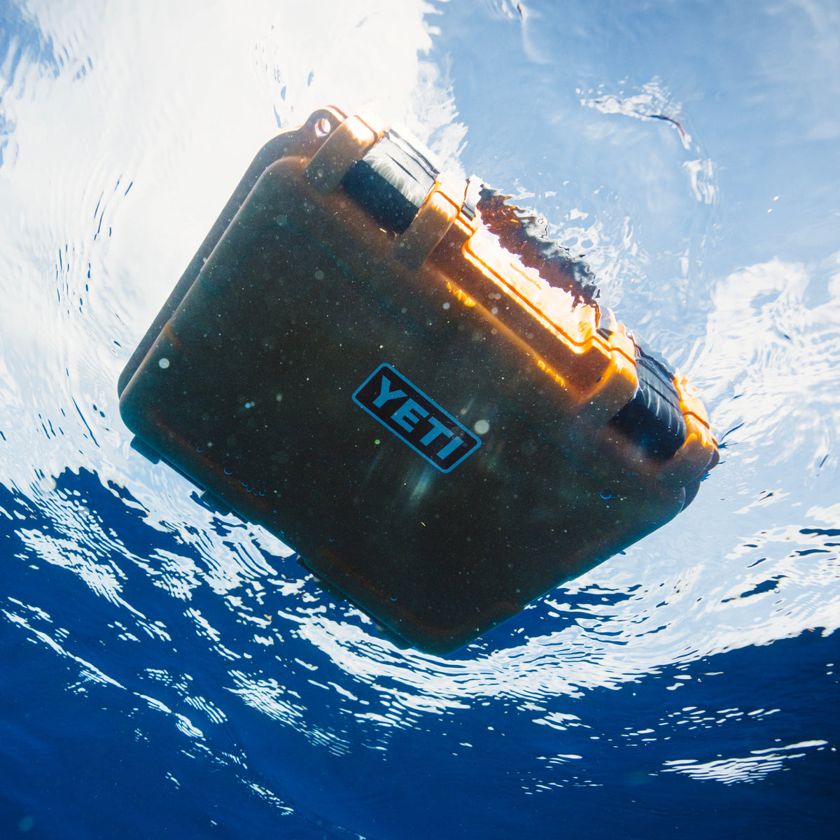 A YETI hard case floating underwater in clear blue ocean water, viewed from below with sunlight reflecting on the water surface.