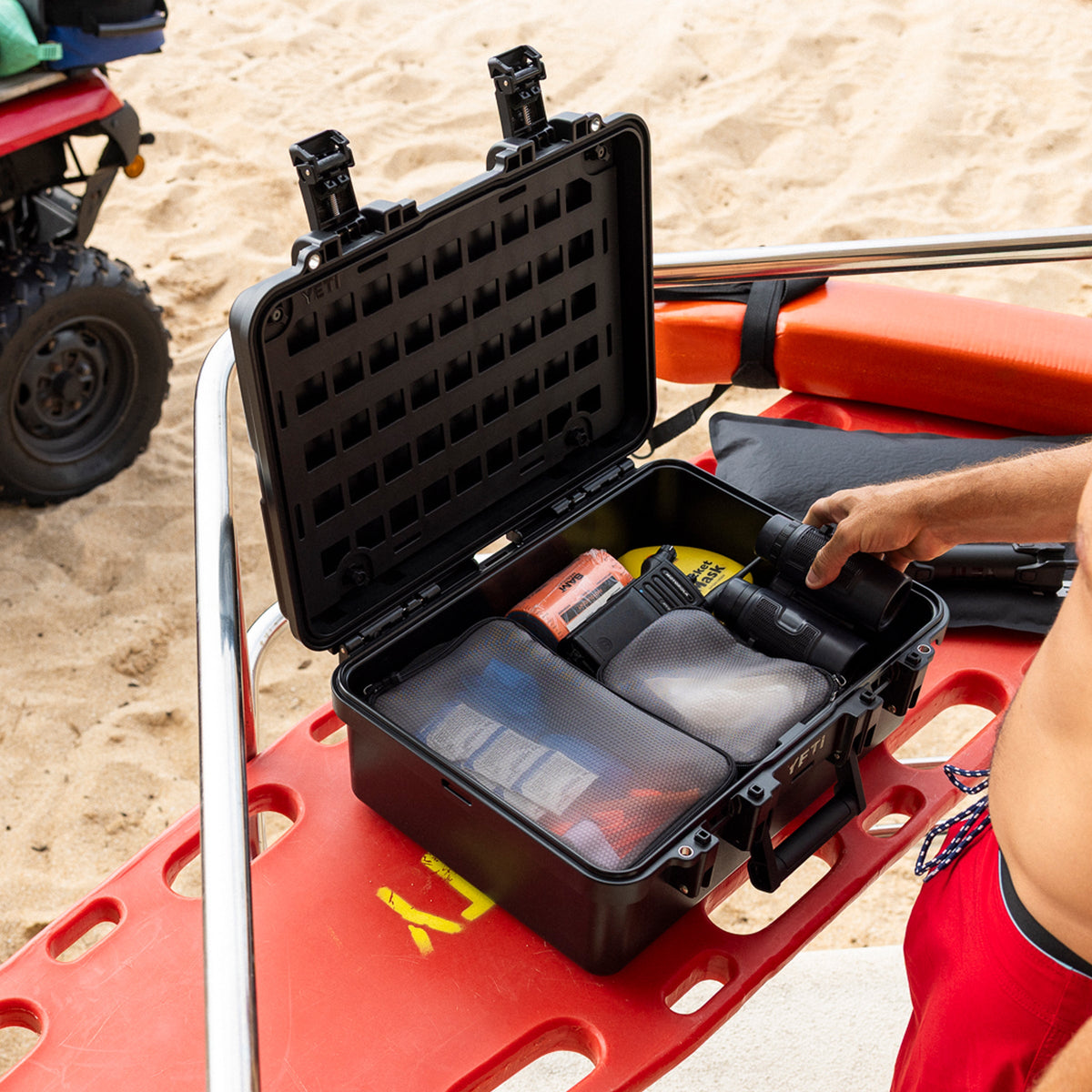 A black hard-shell carrying case opened on a red lifeguard float in a beach setting, with a person's arm reaching inside to retrieve a black flashlight. Sand is visible in the background, along with part of an ATV.