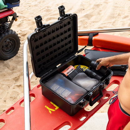 A black hard-shell carrying case opened on a red lifeguard float in a beach setting, with a person's arm reaching inside to retrieve a black flashlight. Sand is visible in the background, along with part of an ATV.