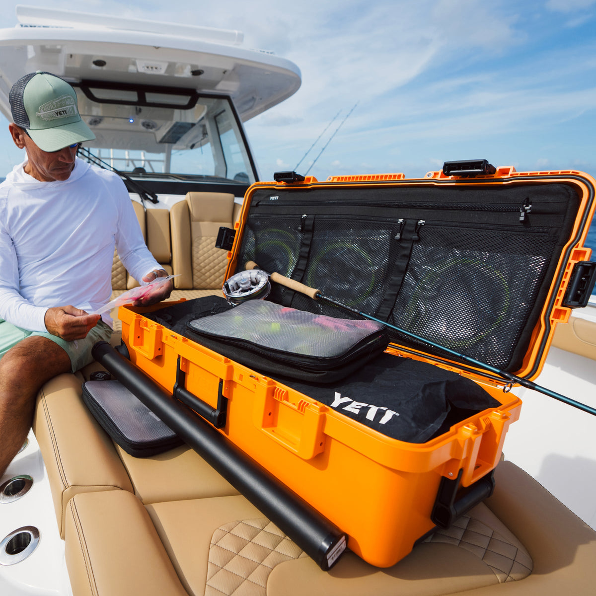 A man sitting on a boat beside an open bright orange YETIT hard case filled with fishing gear, including a black rod case and tackle bags, under a blue sky.