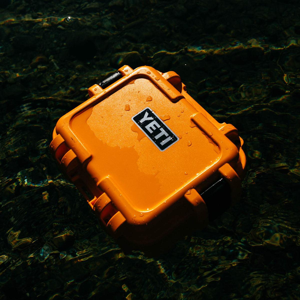 An orange waterproof YETI hard case floating on dark, rippling water with visible ripples and reflections.