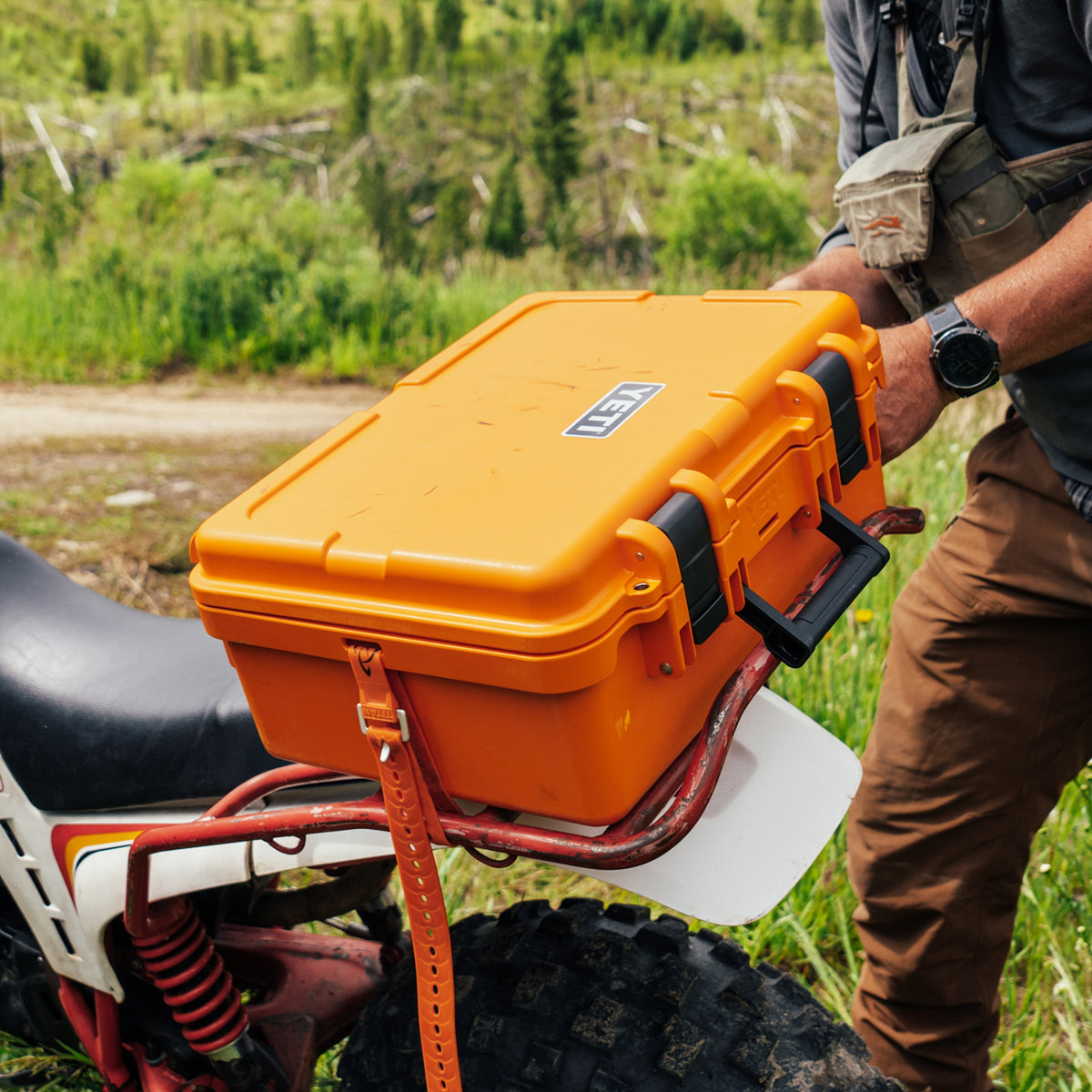 An orange YETI hard case mounted on a red bike rack outdoors on a grassy trail, with a person holding the case and wearing brown pants and a black shirt.