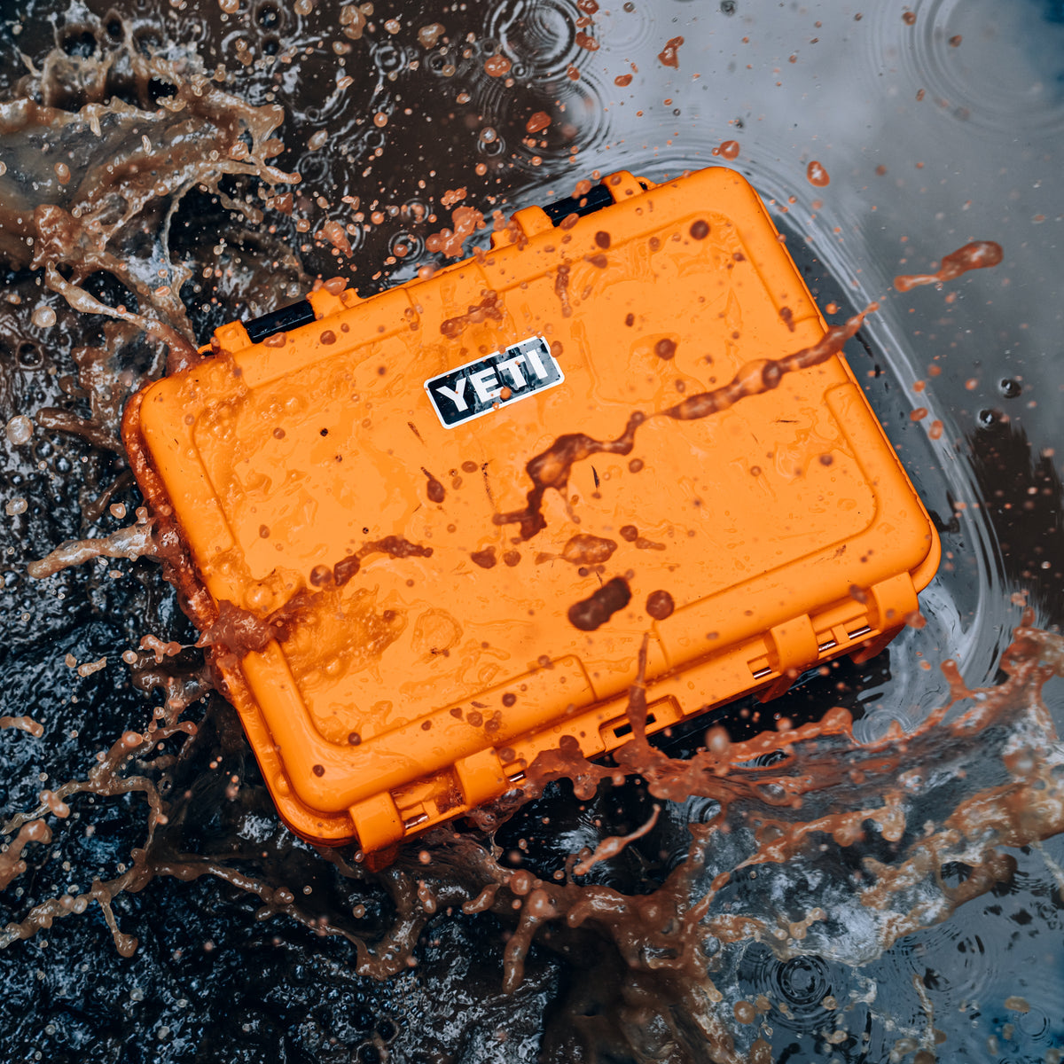 An orange waterproof hard case with a YETI logo, shown splashing through dark water with a brown splash around it.