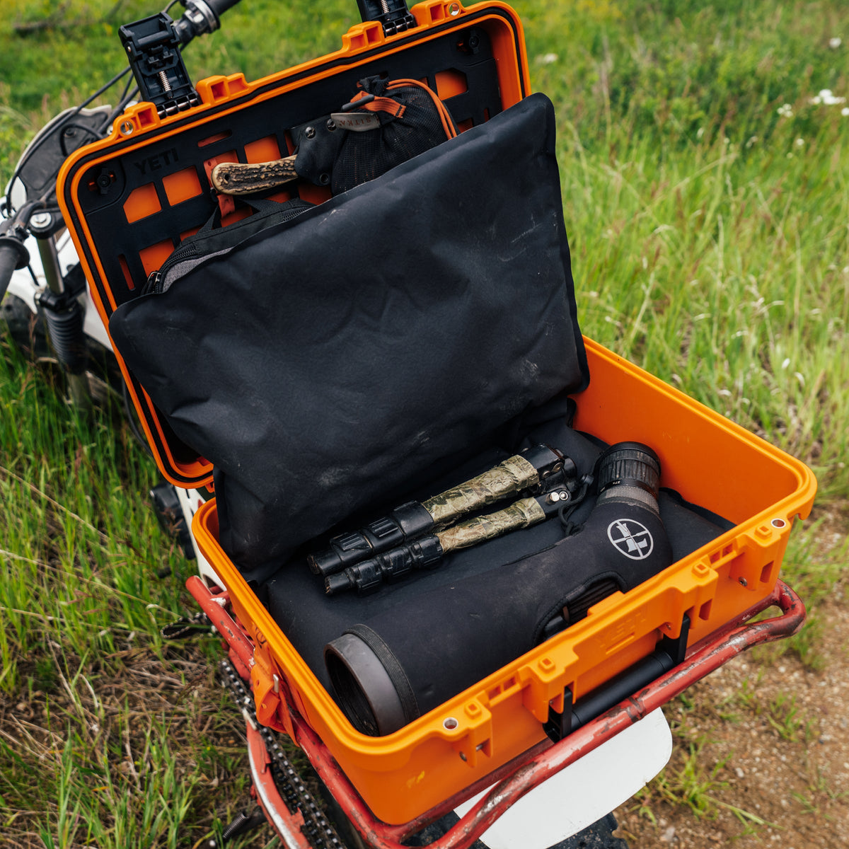 An orange hard-shell case opened on a grassy outdoor surface, containing a black padded organizer with mounted gear including a tripod and binoculars.