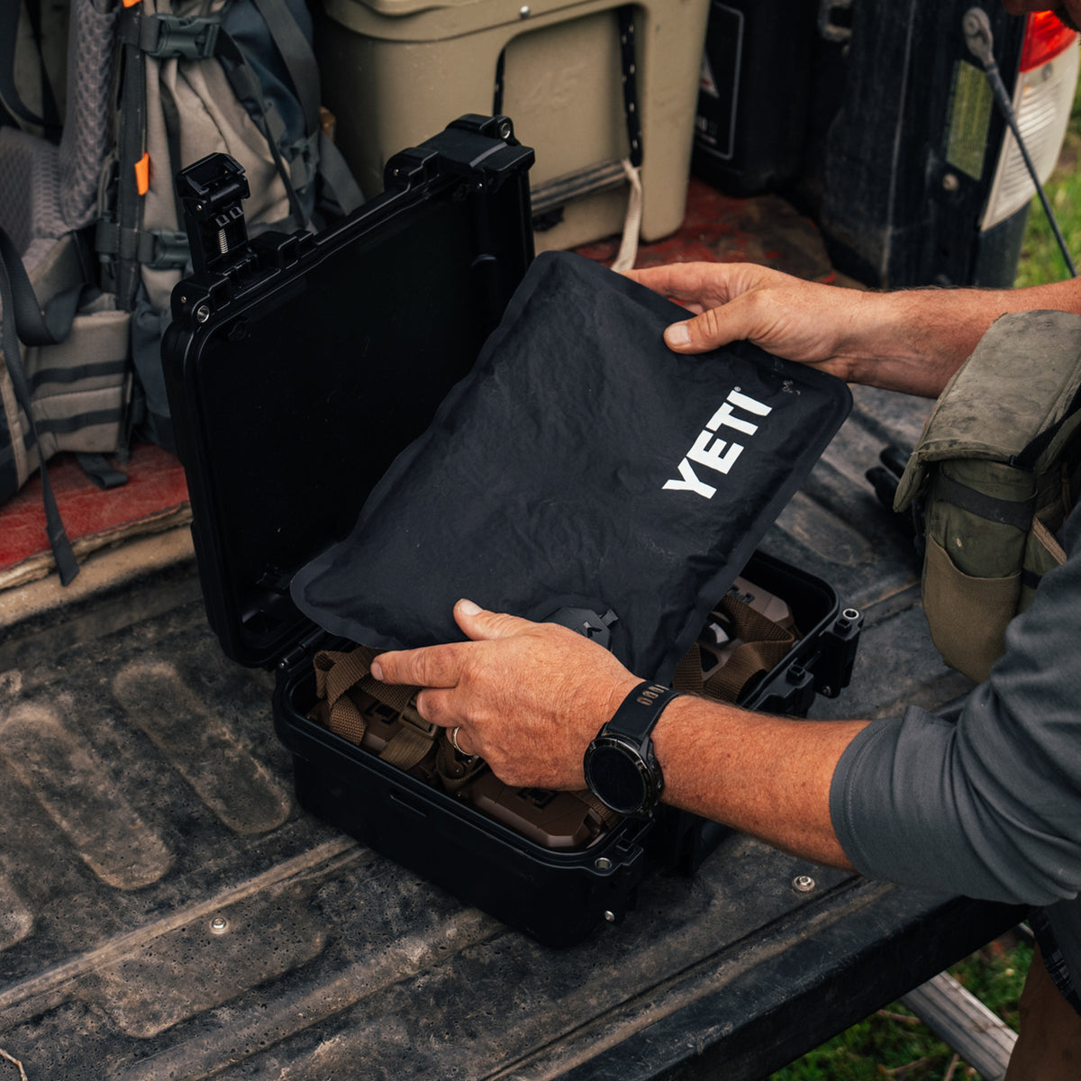 A person's hands open a black YETI hard case and slide a black pouch with the white YETI logo into it, set on the tailgate of a truck with outdoor gear in the background.