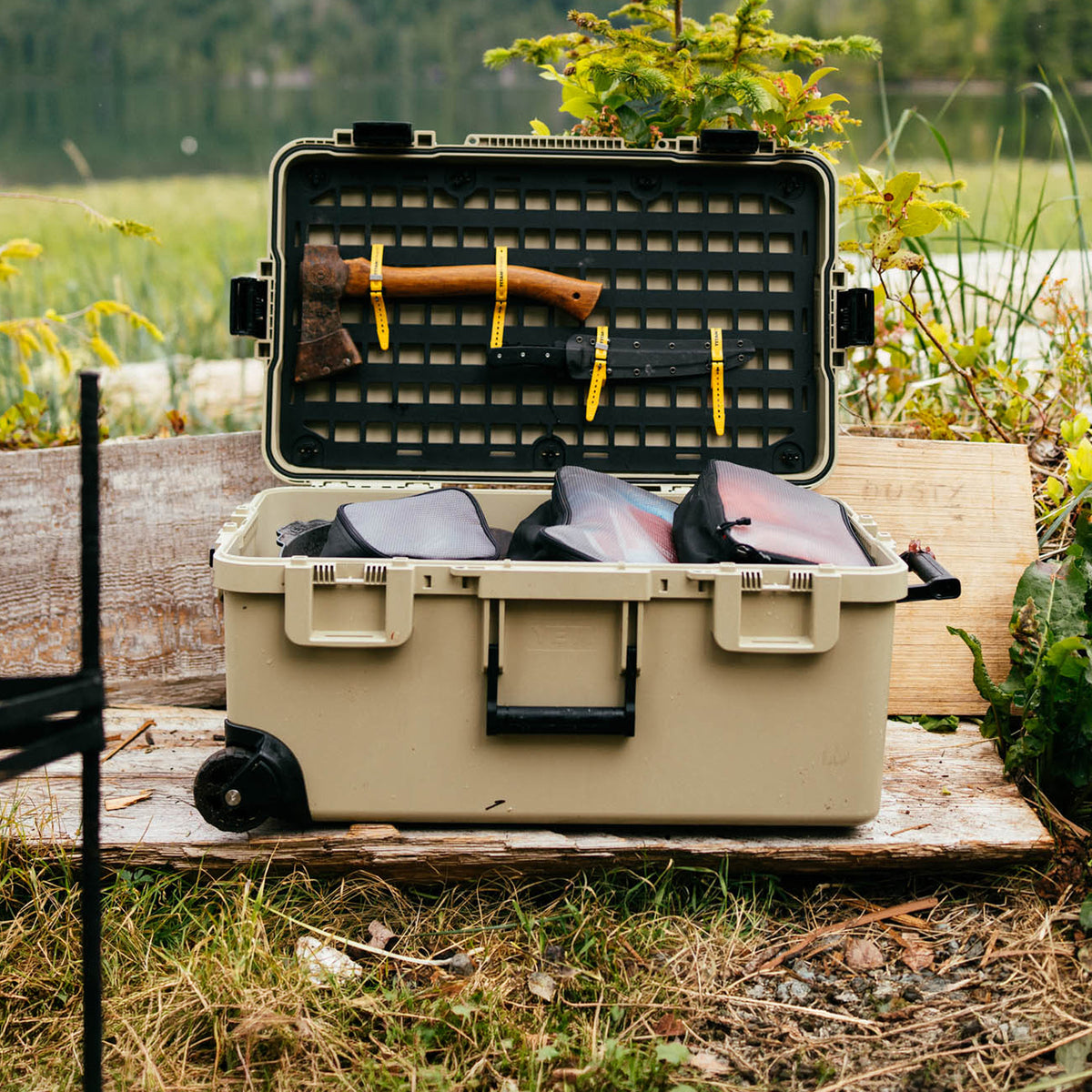 A beige hard-shell outdoor storage case with wheels is open on a wooden platform in a grassy outdoor setting. The case is shown with its lid open, revealing a black foam organizer insert holding an axe and a black rifle secured with yellow straps, along with several gear pouches inside.