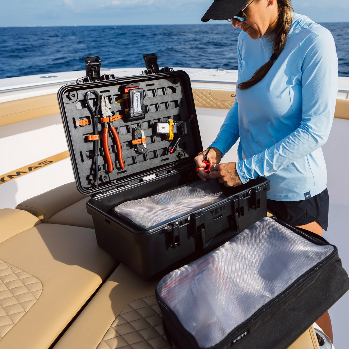 A woman in a light blue long-sleeve shirt and black cap organizes tools inside an open black hard carrying case on a boat deck, with the matching zippered bag placed beside it. The ocean and boat are visible in the background.