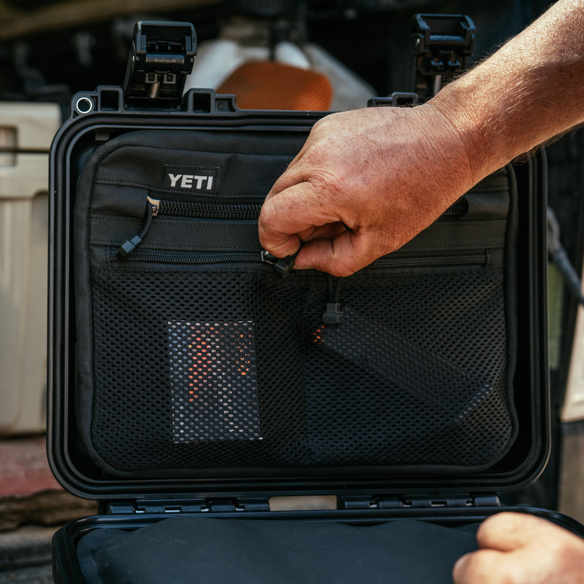 A close-up of a person's hand organizing gear inside an open black YETI hard case, showing the interior mesh pockets and zipper compartments.
