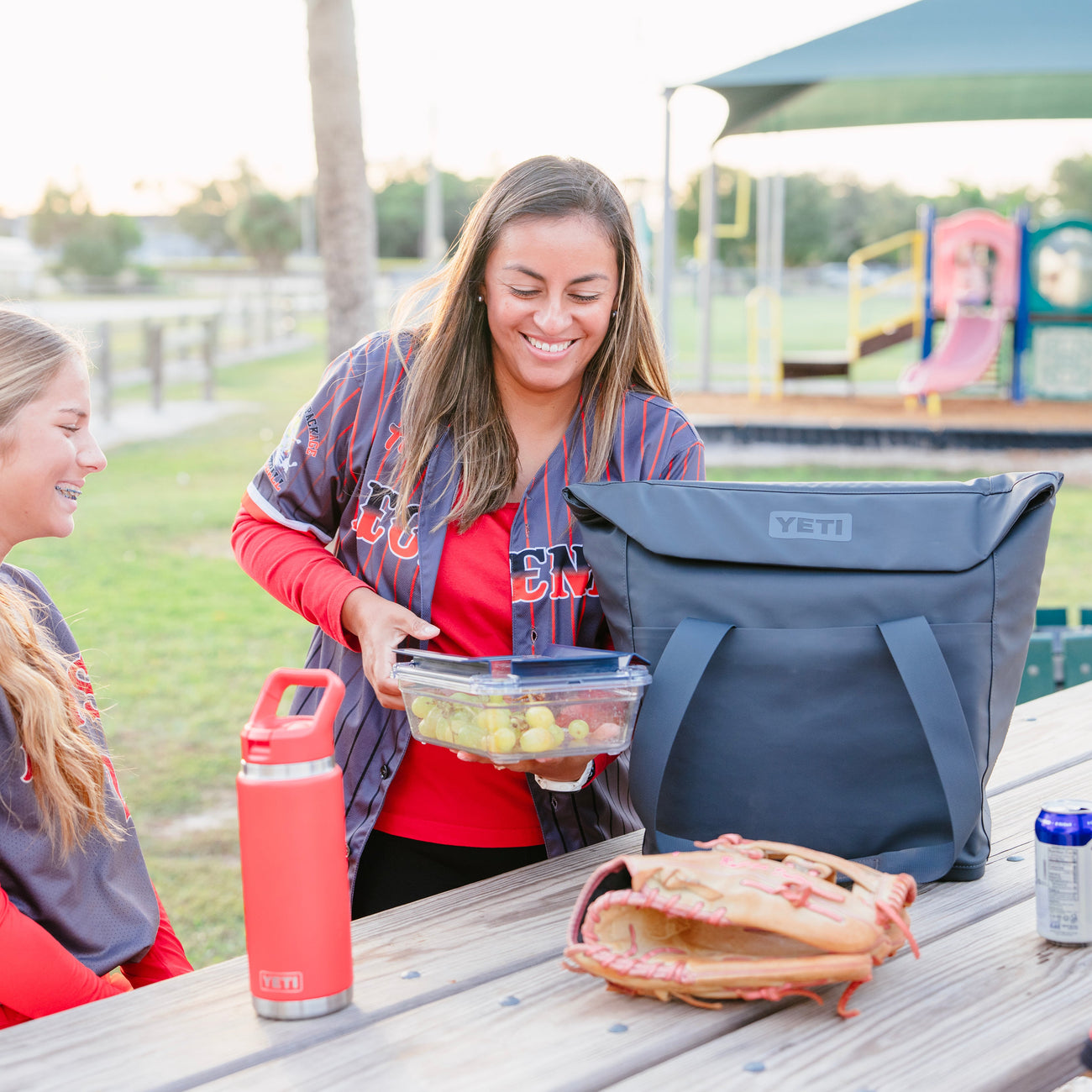 Two women and a child at a picnic table with a YETI cooler and sports equipment.